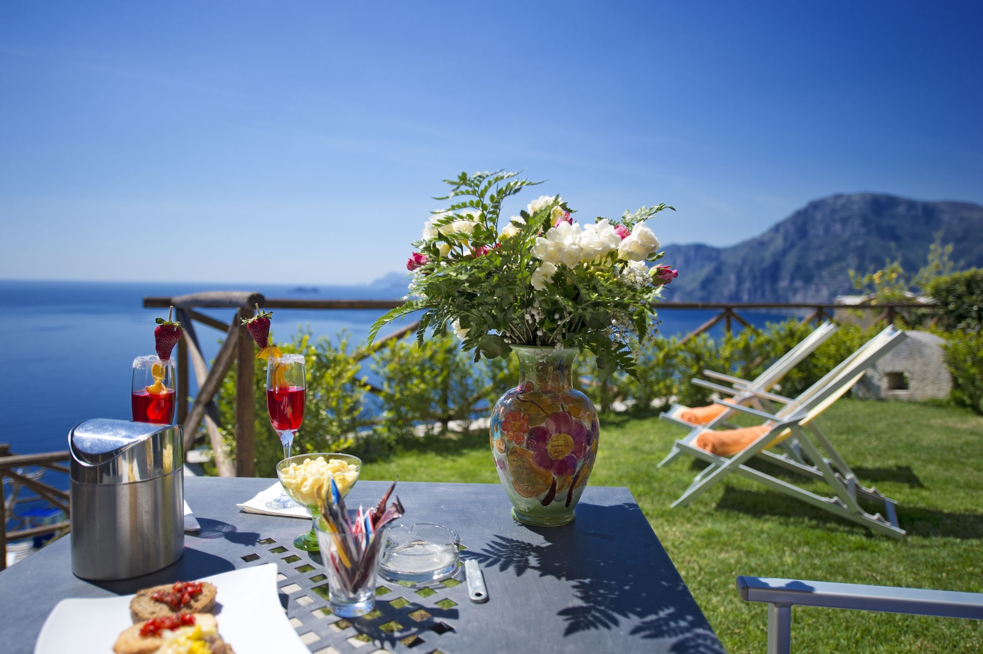 Una mesa con un jarrón de flores, una jofaina y un vaso de bebida roja, frente a una vista panorámica del mar y las montañas.