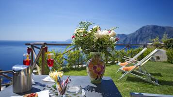 Una mesa con un jarrón de flores, una jofaina y un vaso de bebida roja, frente a una vista panorámica del mar y las montañas.