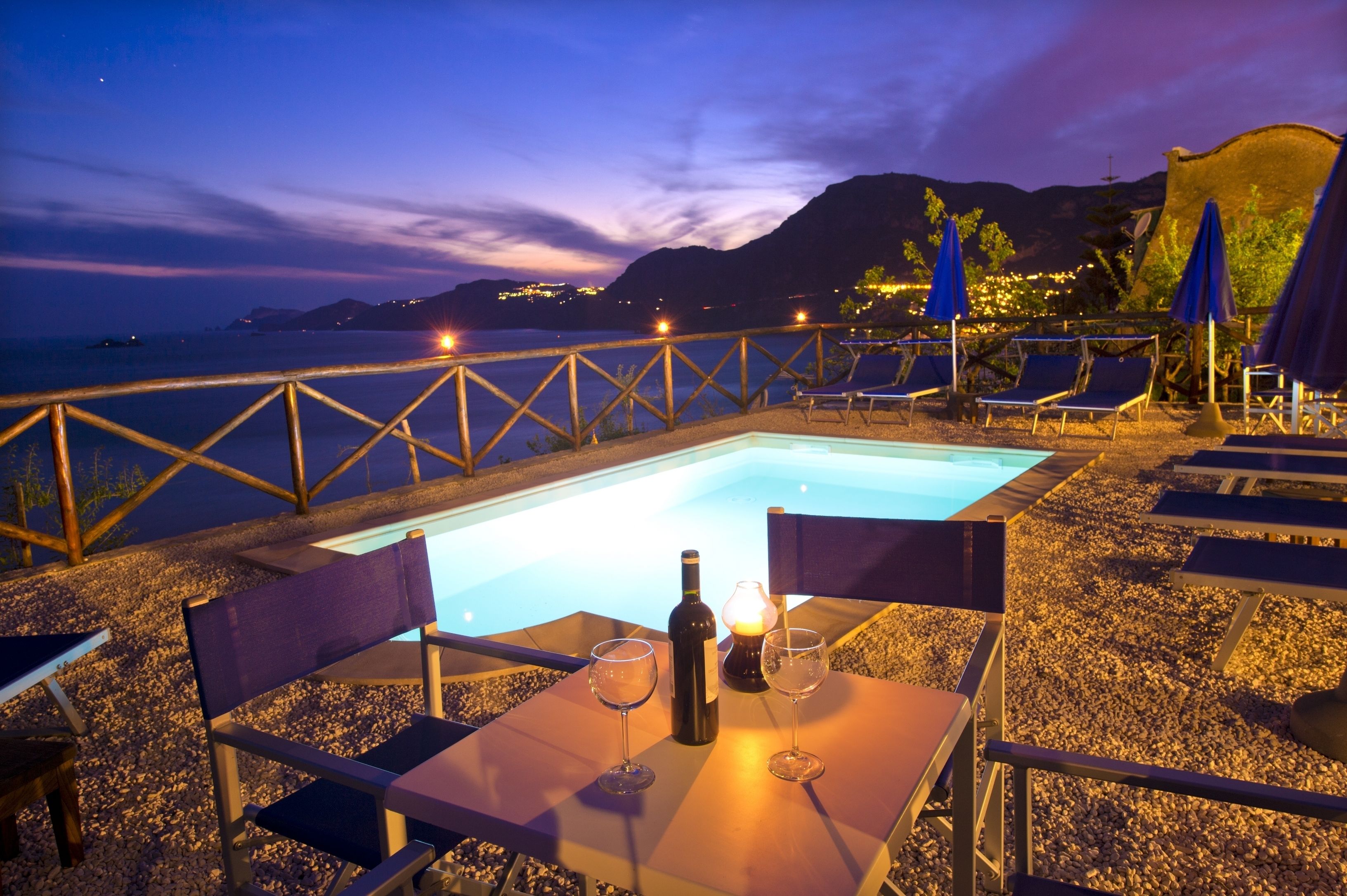 An outdoor pool area with lounge chairs and a table, overlooking a body of water and hills at dusk.
