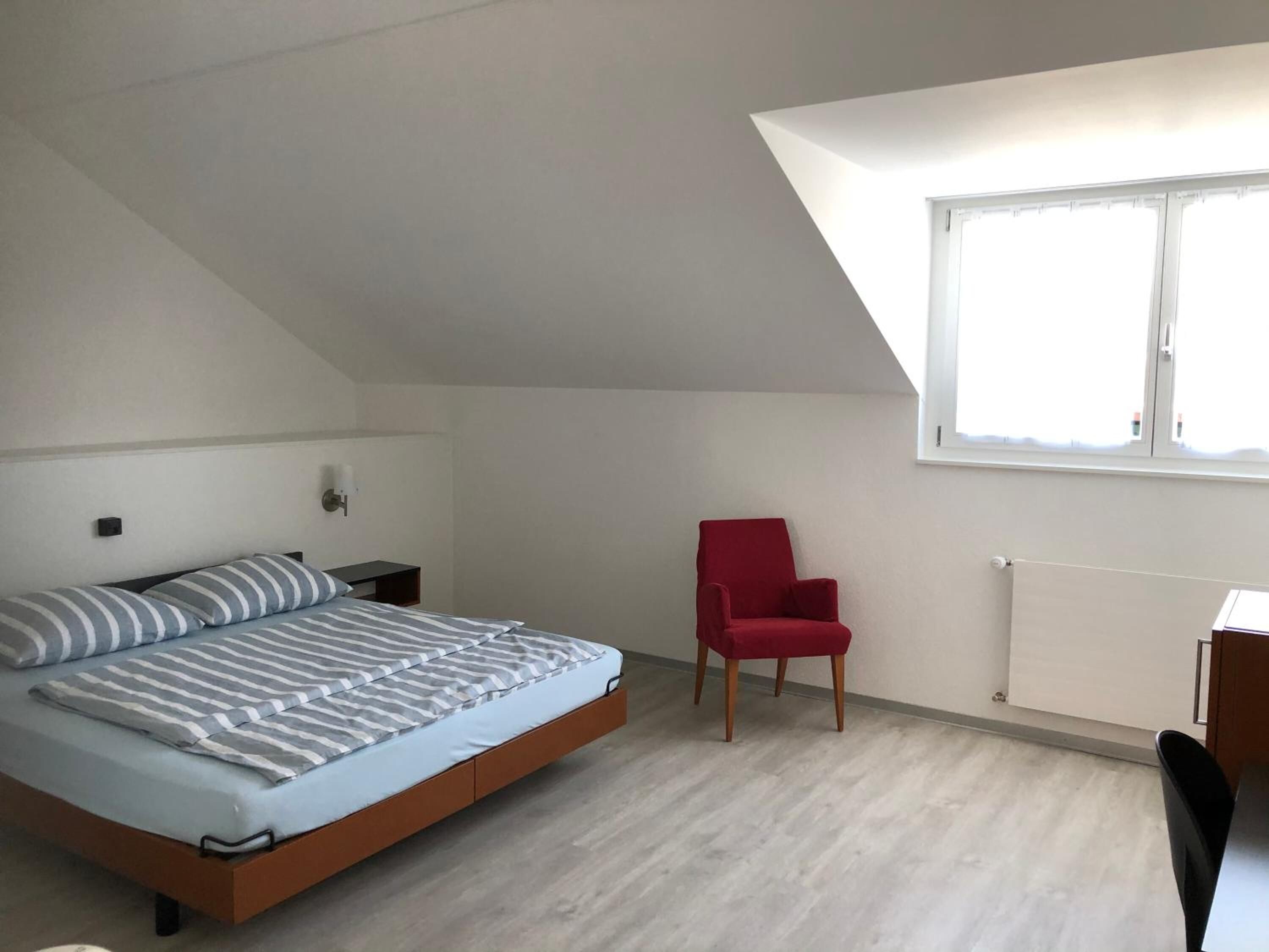A bedroom with a sloped ceiling, a bed with striped bedding, a red chair, and a window providing natural light.