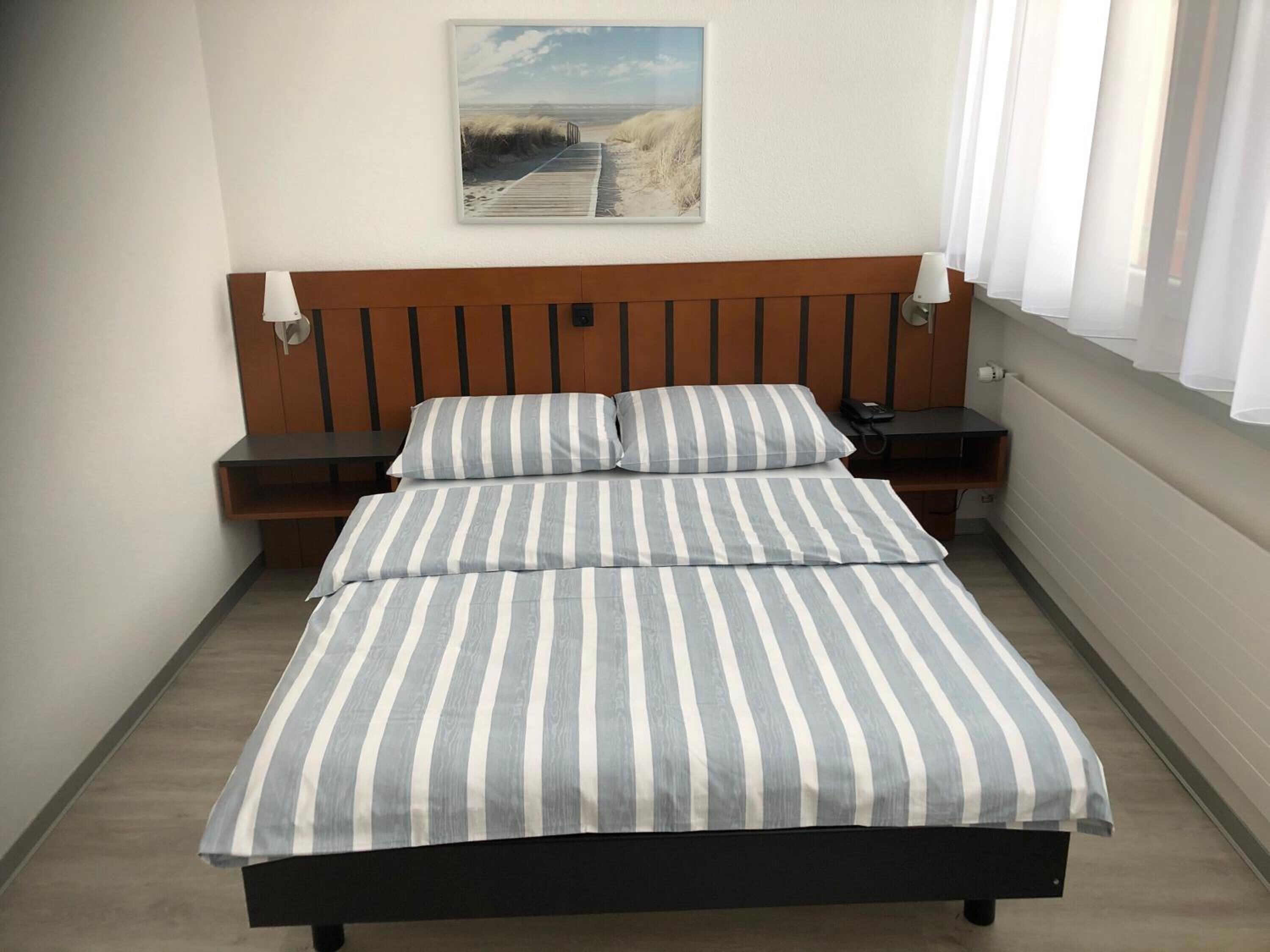 Un dormitorio con cabecera de madera, cama con ropa de cama a rayas, una mesita de noche, una lámpara y un cuadro en la pared.