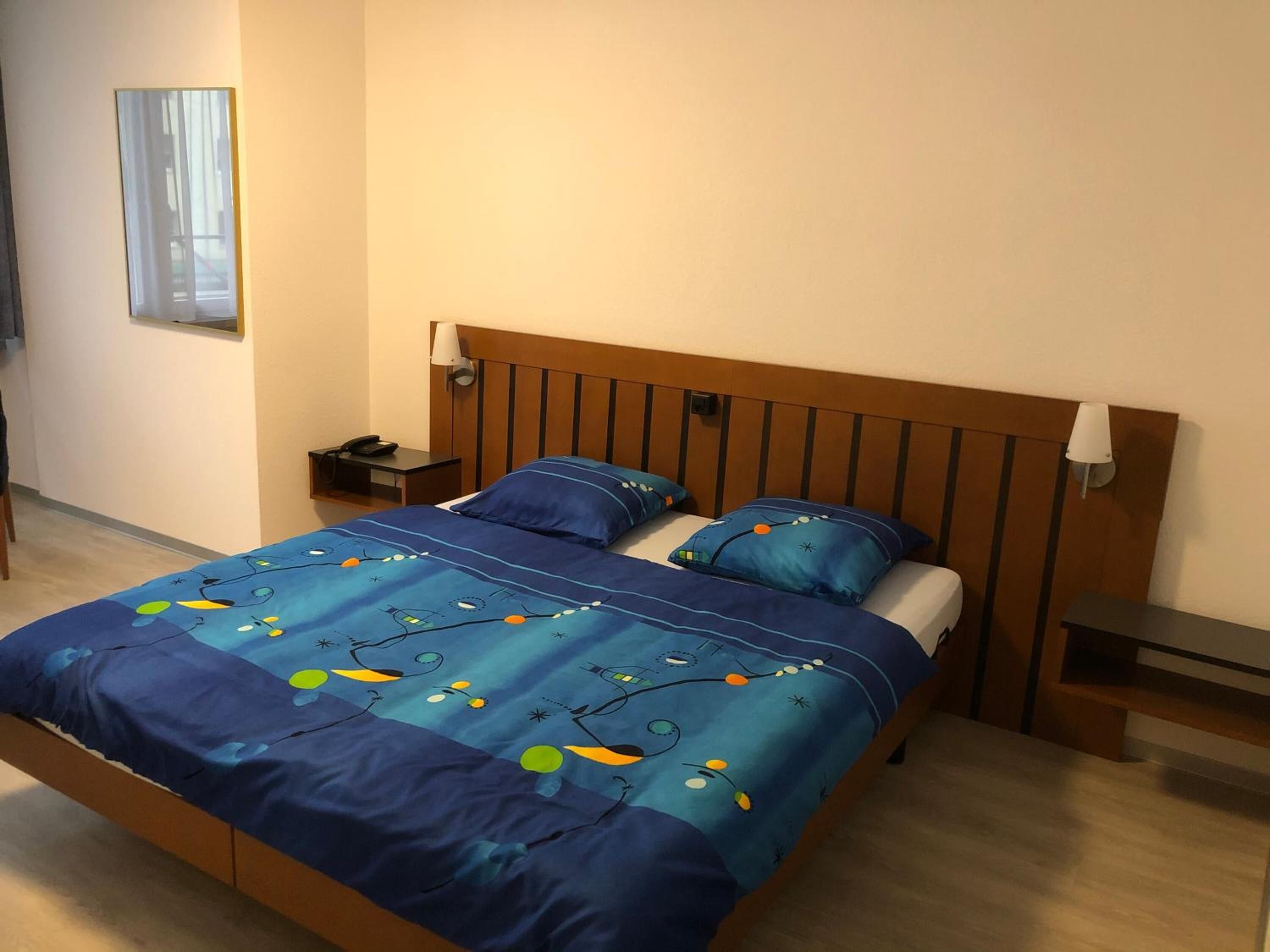 Un dormitori amb un panell de fusta al capçal, dues taules de nit, una cambrera i un llit amb una col·cha i almoines blaves amb estampat floral.