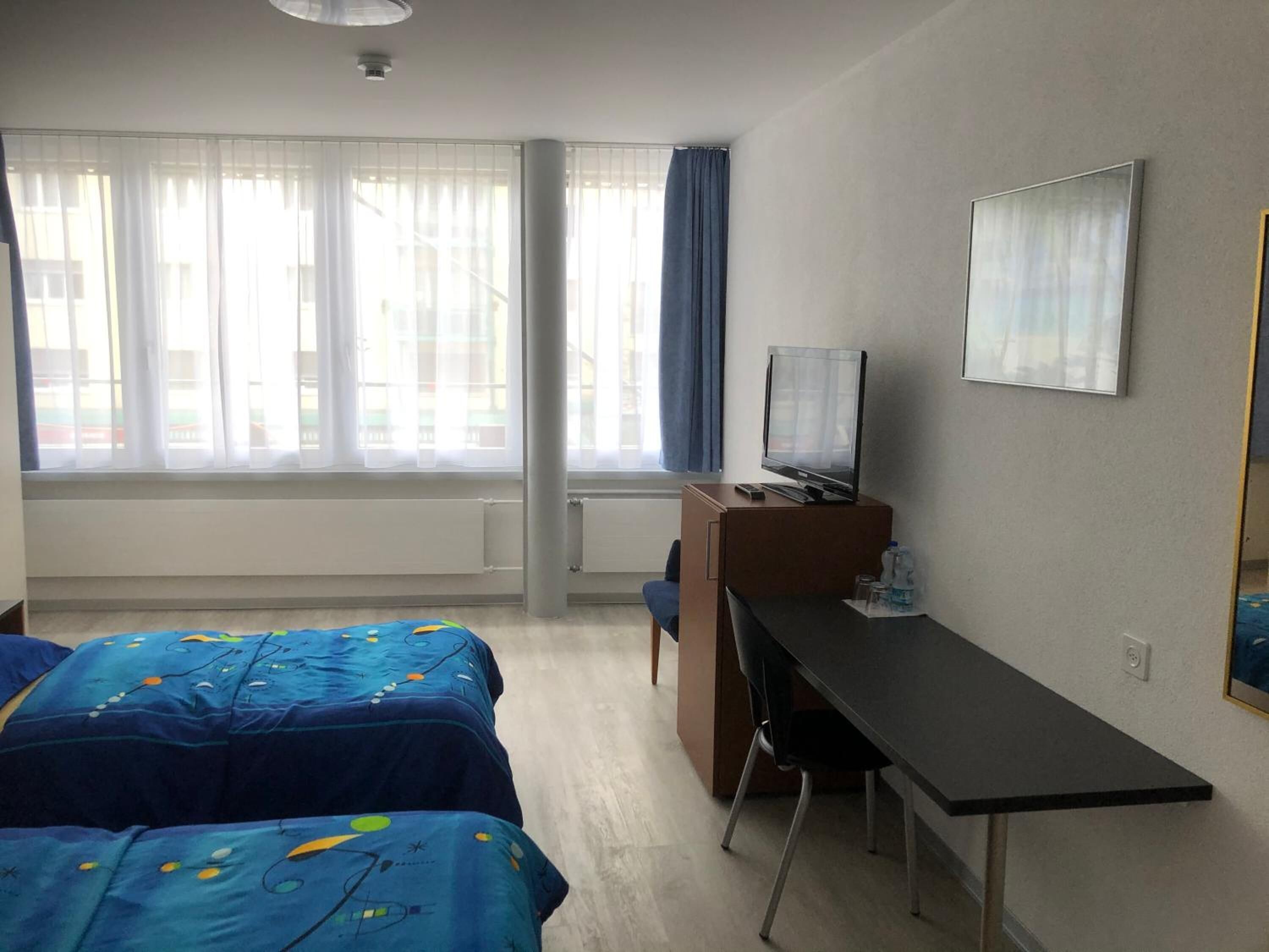 Ein Hotelzimmer mit zwei Betten, einem Schreibtisch, einem Fernseher und einem Fenster mit Vorhängen.