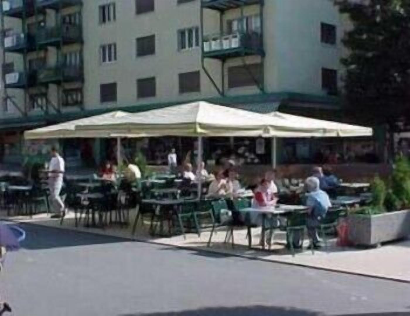 Un café en plein air avec des tables et des chaises, un bâtiment avec des balcons en arrière-plan, et des personnes qui déjeunent ou dînent.