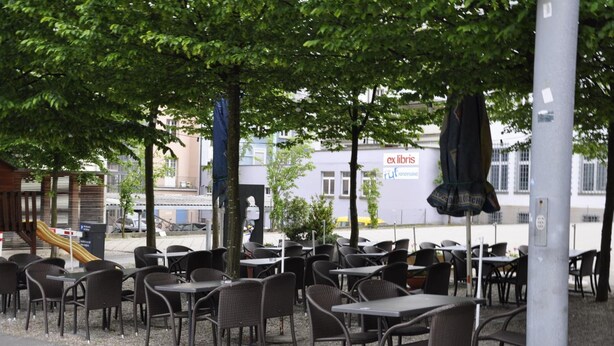 Outdoor seating with tables and chairs, a playground, and a building with signage in the background.
