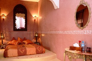 A bedroom with a bed, two wall sconces, a mirror, and a bedside table with a tissue box, a bottle, and a vase with a rose.