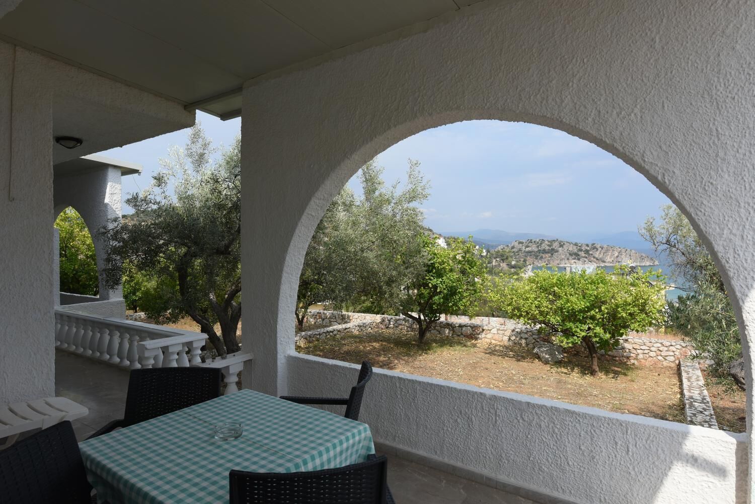 Un espacio de estar al aire libre cubierto, con una mesa y sillas, vista a olivos y un paisaje lejano.