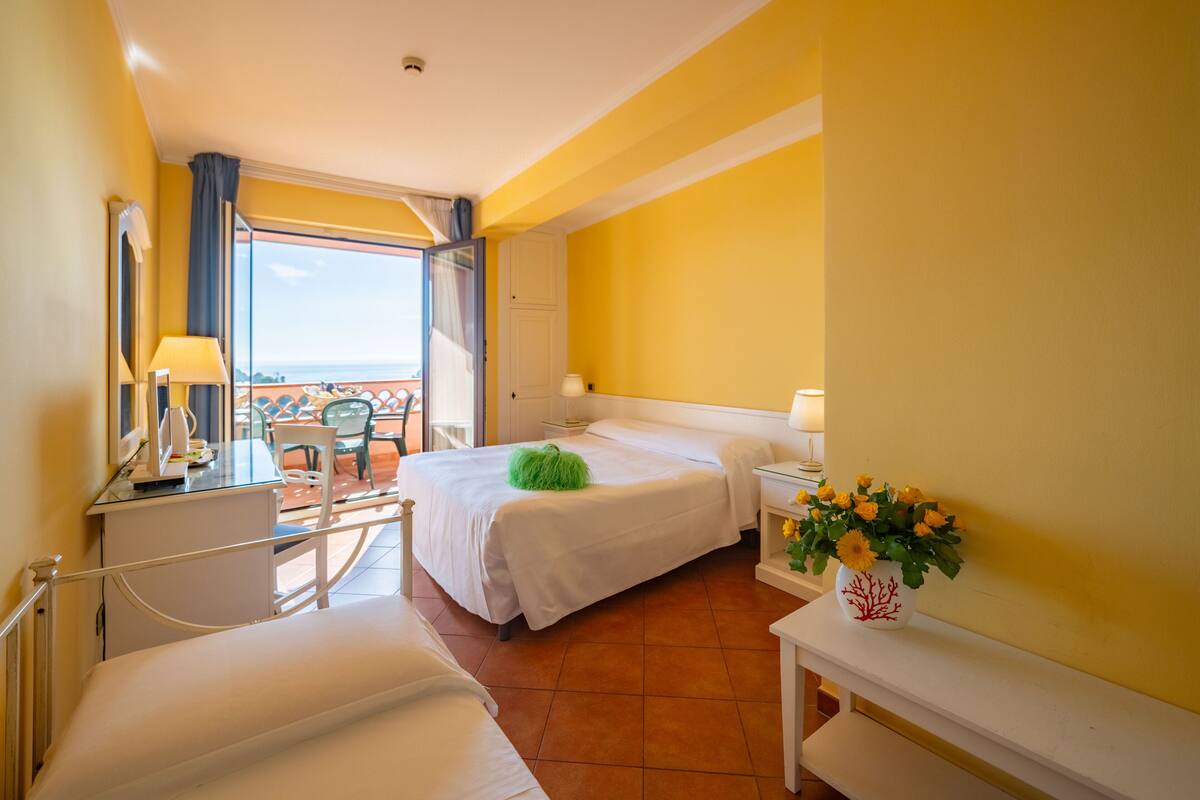 A hotel room with a bed, a desk, a chair, a small table with a vase of flowers, and a balcony with a view.