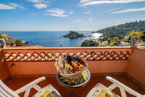 A balcony with a table set for breakfast, overlooking a coastal view with a rocky island and buildings. - Mendolia Beach Hotel (Taormina)