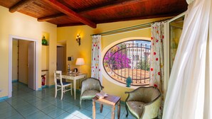 Una habitación acogedora con techo de madera, un ventanal redondo con cortinas florales, dos sillas, una mesita con un libro y una lámpara.
