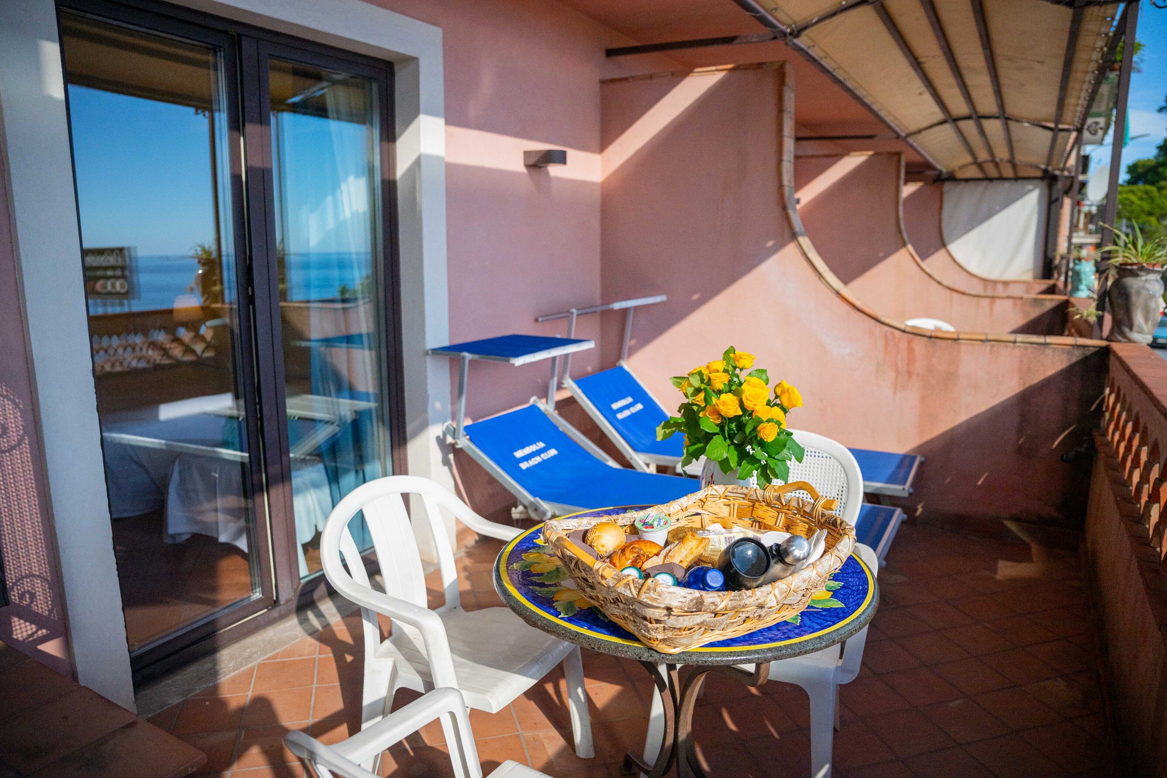 Un balcon avec une table dressée pour deux personnes, un panier en osier rempli de fruits et un vase de fleurs, et une vue sur la mer par une porte vitrée.