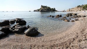 Una playa de guijarros con agua cristalina, afloramientos rocosos y una isla distante con edificios.