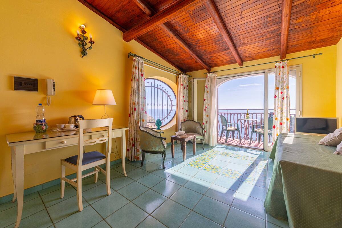 A room with a wooden ceiling, a desk with a chair, a bed, and a balcony with a view.