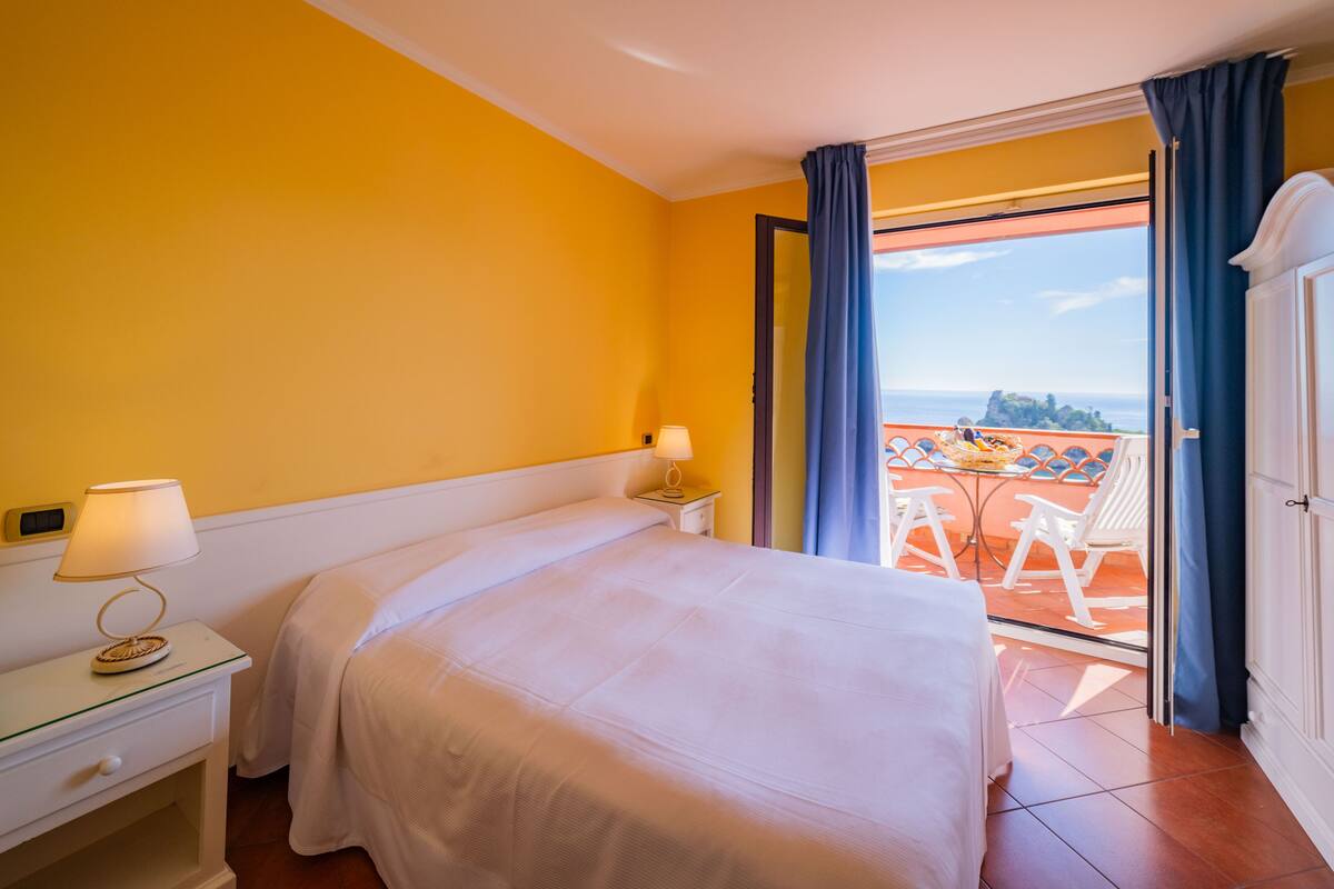 A hotel room with a bed, a nightstand, a lamp, and a view of an outdoor terrace with chairs.