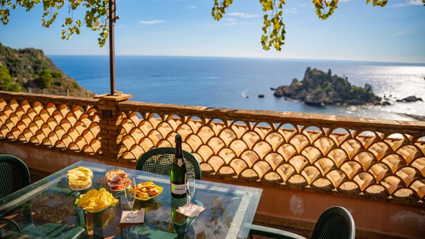 Una mesa con una botella de vino, copas y comida en una terraza con vistas al mar.