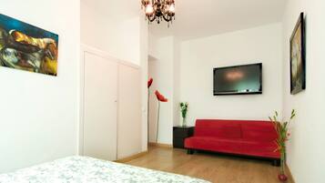 Un dormitorio con cama, un sofá rojo, televisor, candelabro y obras de arte en las paredes.