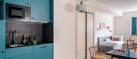 A modern kitchen with teal cabinets, a microwave, and a sink. There is a dining area with a table and chairs, and a bed in the background.