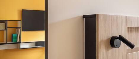 Una habitación de hotel moderna con cabecera de madera, televisor de pantalla plana montado en la pared, mesita de noche con lámpara y sofá con cojín.