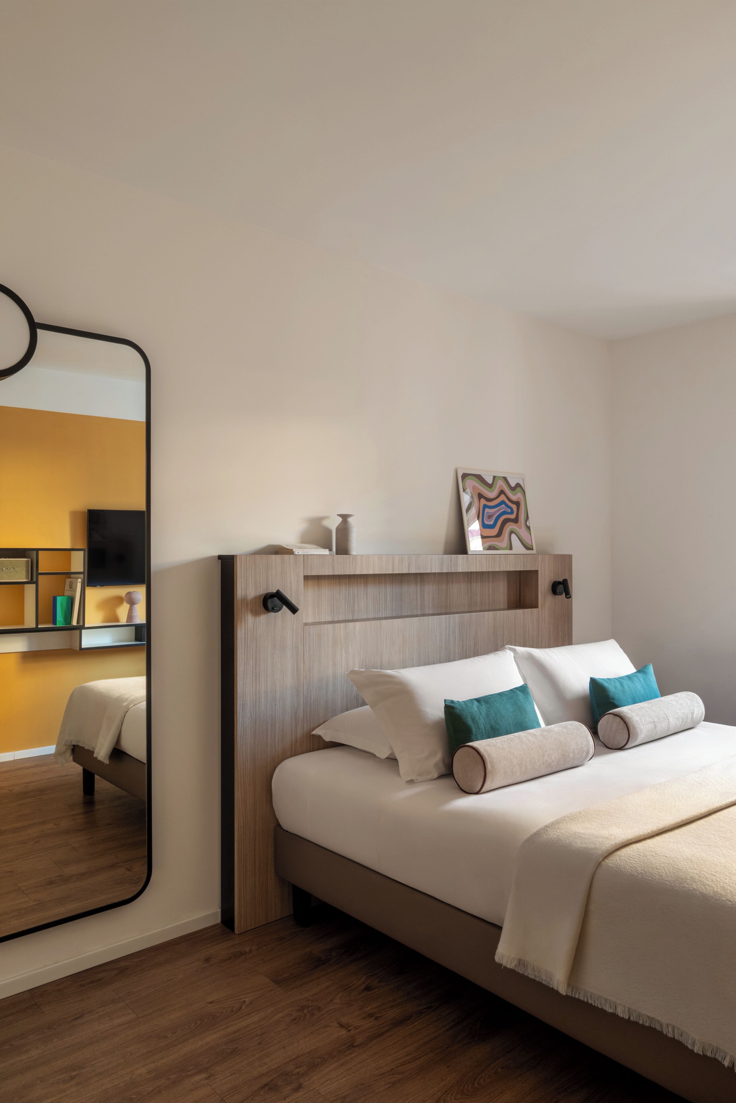 Une chambre d’hôtel moderne avec une tête de lit en bois, un grand miroir, un lit avec du linge de lit blanc et des coussins turquoise, ainsi qu’une petite étagère ornée d’objets décoratifs.