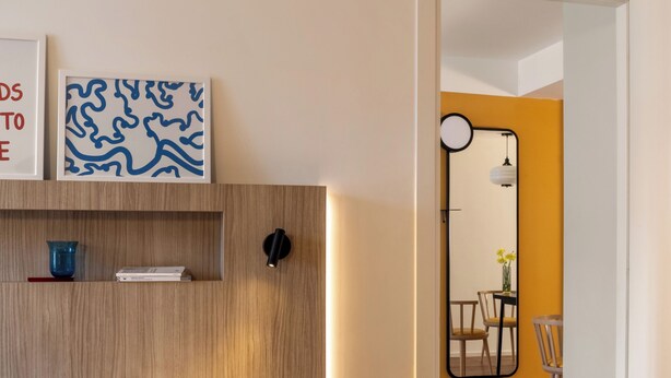 Habitación de hotel moderna con cabecera de madera, cama con ropa de cama blanca y marrón, un pequeño estante con adornos, un espejo con marco redondo y un área de comedor con pared amarilla y suelo de madera.