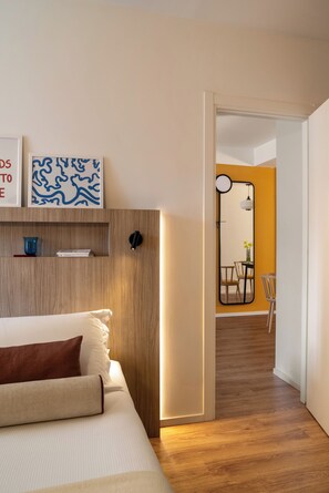 Habitación de hotel moderna con cabecera de madera, cama con ropa de cama blanca y marrón, un pequeño estante con adornos, un espejo con marco redondo y un área de comedor con pared amarilla y suelo de madera.