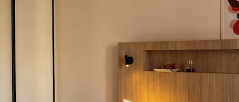 A hotel room with a bed, pillows, a nightstand, a wall-mounted shelf, and a framed picture.