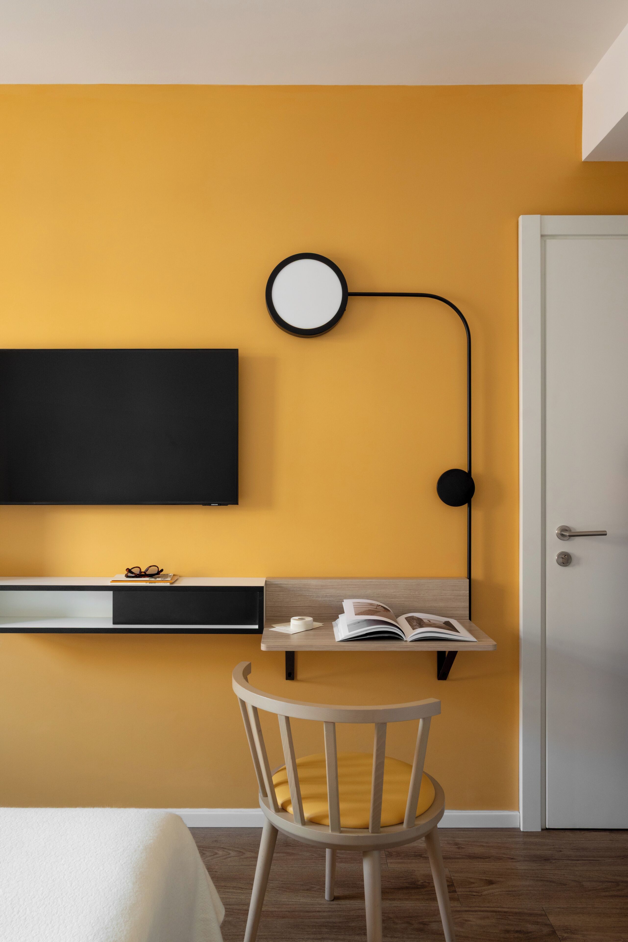 Habitación moderna con una pared amarilla, un televisor de pantalla plana montado en la pared, un escritorio con una silla y una puerta.