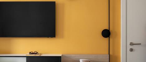 A modern room with a yellow wall, a flat-screen TV mounted on the wall, a desk with a chair, and a door.