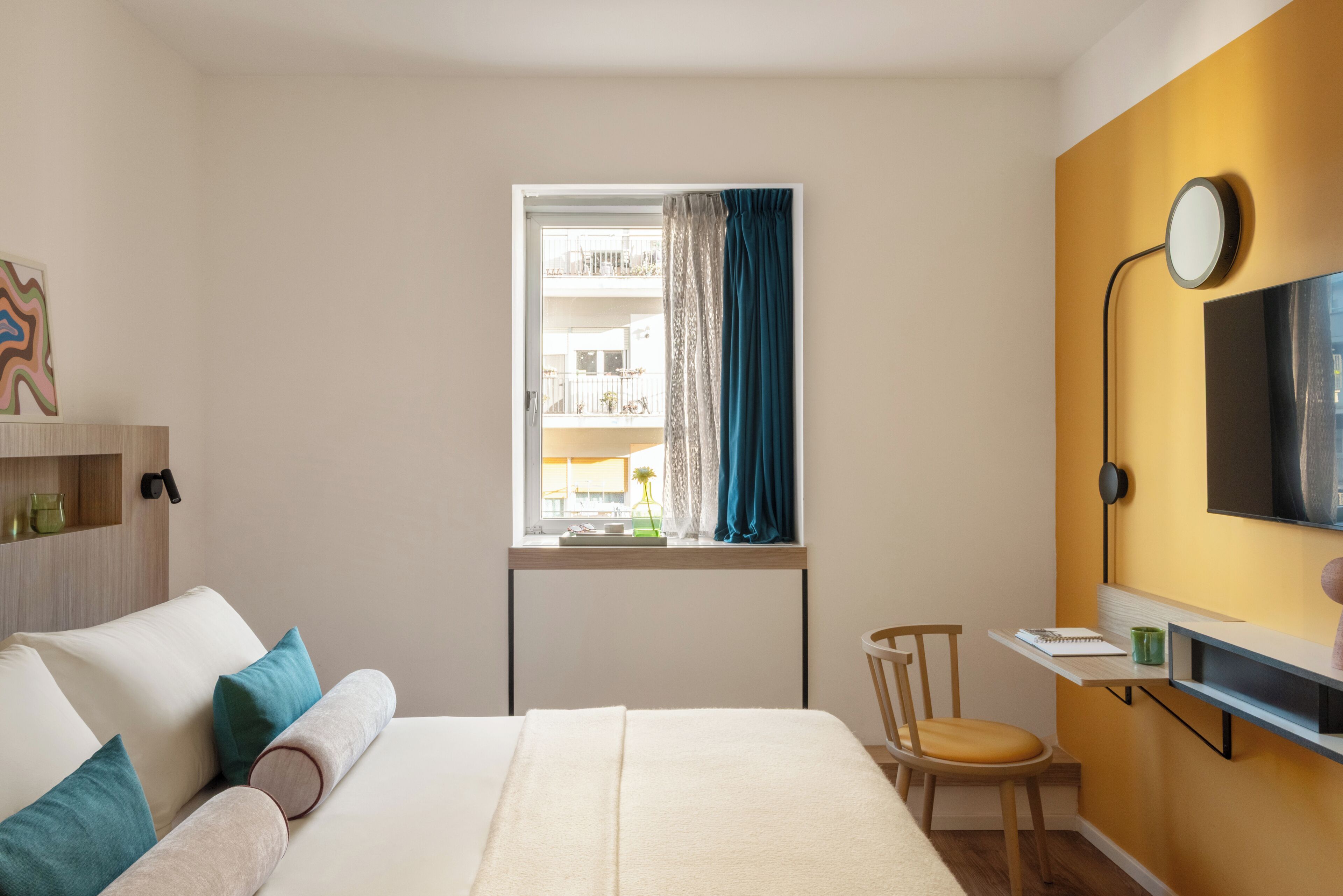A bedroom with a bed, a desk, a chair, a window with curtains, a TV, and a shelf with items.