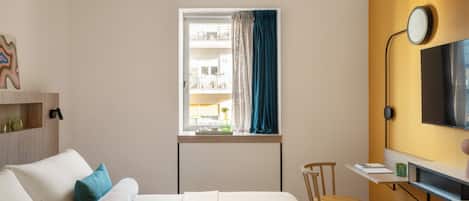 A bedroom with a bed, a desk, a chair, a window with curtains, a TV, and a shelf with items.