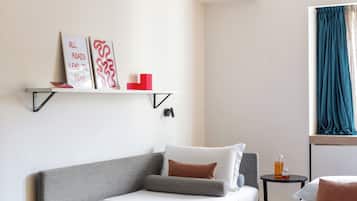 Habitación de hotel con dos camas, una mesita, una estantería con obras de arte y una ventana con cortinas.