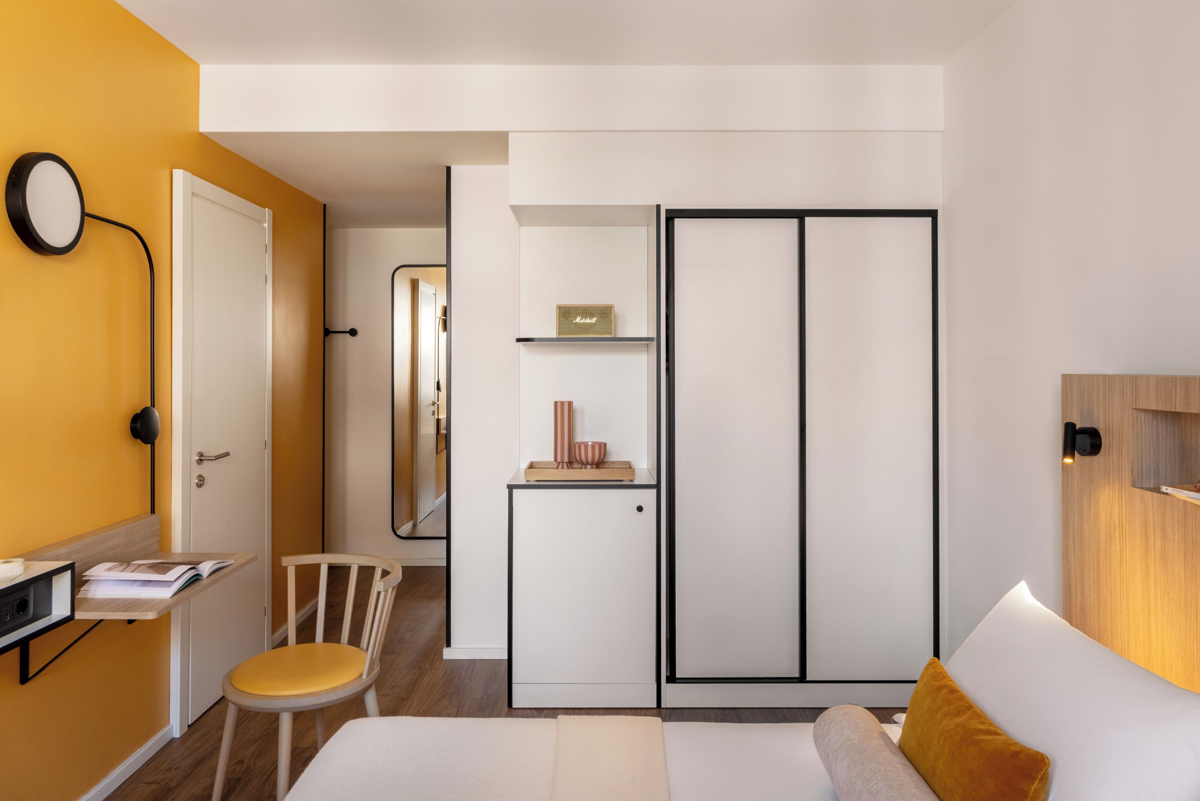 A modern bedroom with a bed, a desk with a chair, a wardrobe, and a bedside table.