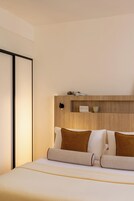 Una camera d'albergo moderna con un letto, cuscini, un comodino e una mensola a parete con una fotocamera e una tazza.
