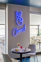 Un neon con un simbolo di pace e la scritta 'Cheers!' sopra un tavolo con una bevanda e un piatto di cibo.