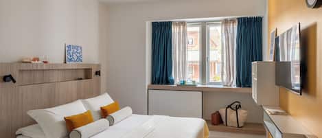Habitación de hotel con cama, escritorio, silla, televisor, ventana con cortinas y estante con objetos.