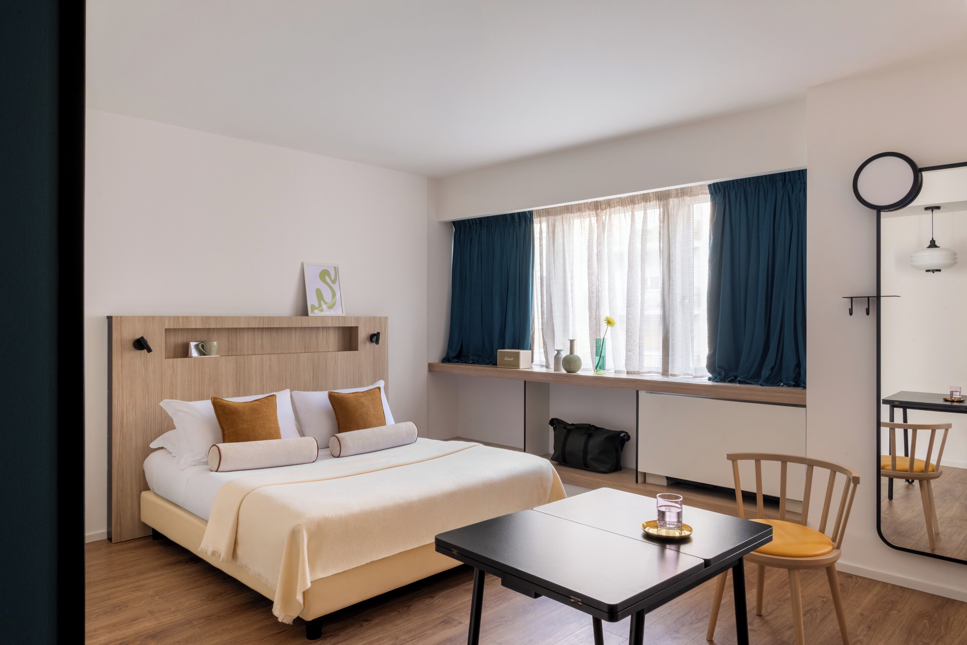 A hotel room with a bed, a desk, a chair, a window with curtains, and a mirror.