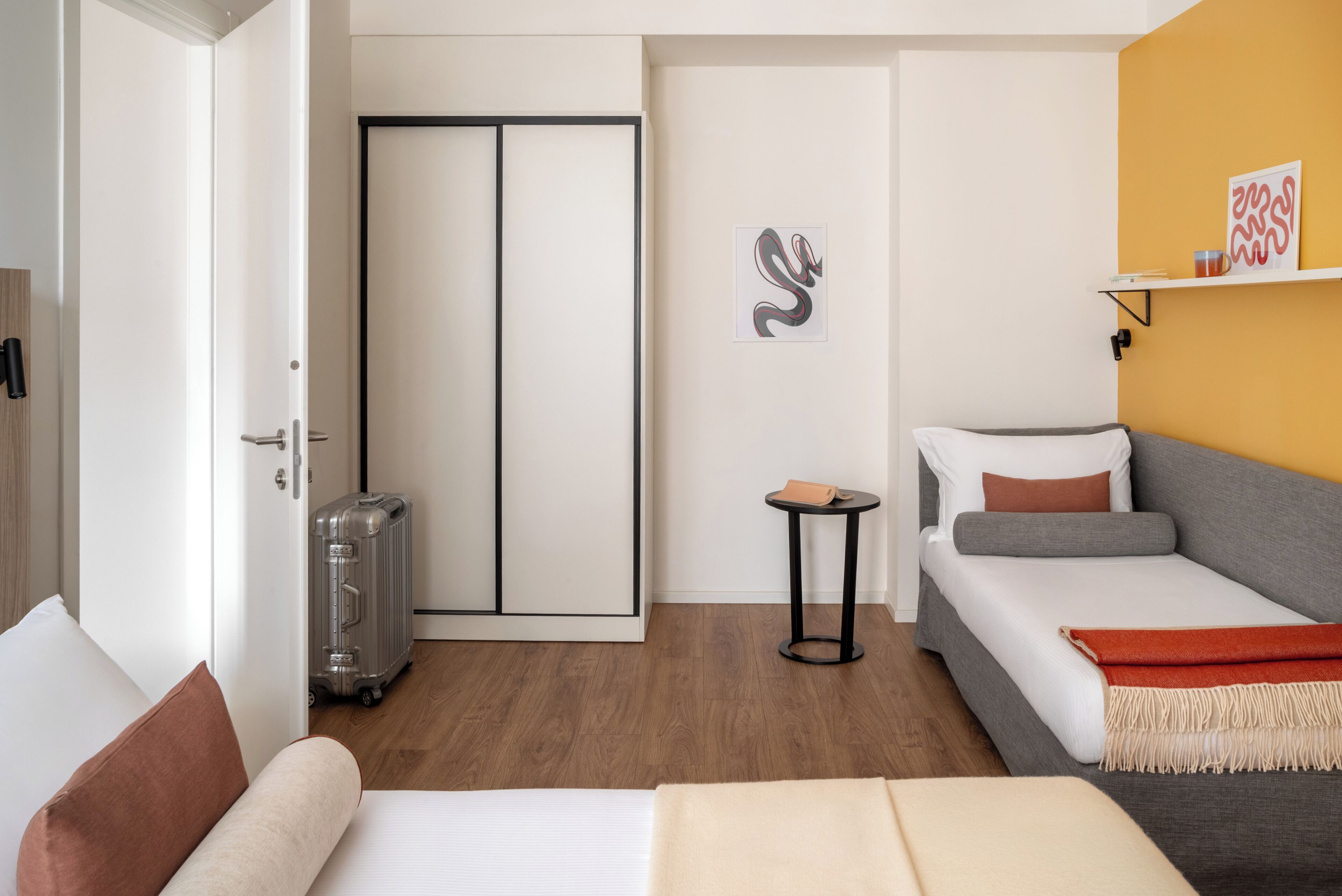 Une chambre d’hôtel avec un lit, une petite table, une valise, une armoire et un mur orné d’un tableau.