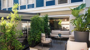 Un espacio exterior moderno con zona de estar que incluye un sofá, sillas y una mesa de centro, rodeado de vegetación y vistas a un edificio.