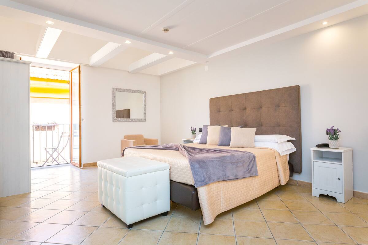 A modern bedroom with a large bed, a white nightstand, a white chair, and a sliding glass door leading to an outdoor area.