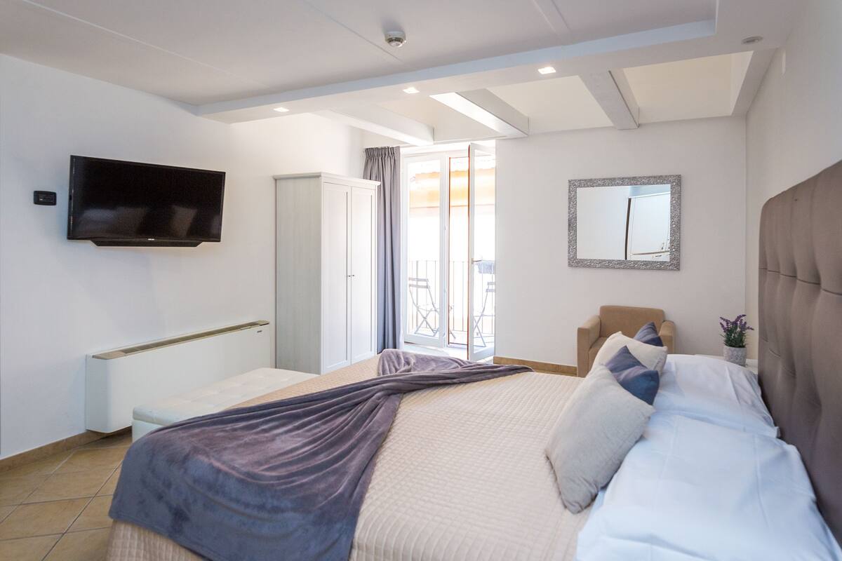 A bedroom with a bed, a TV mounted on the wall, a chair, a small table, and a balcony with a view.