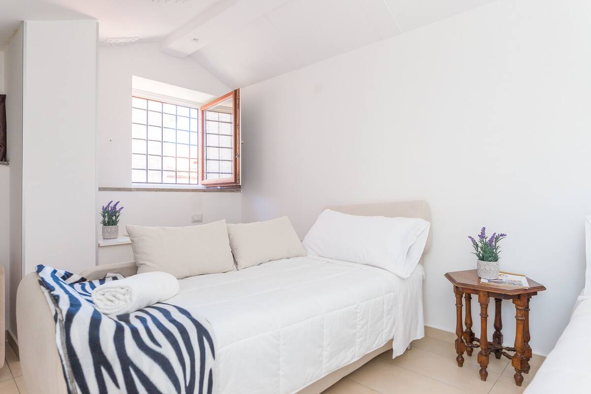 A bedroom with a bed, a window, a small table with a plant, and a shelf with a plant.