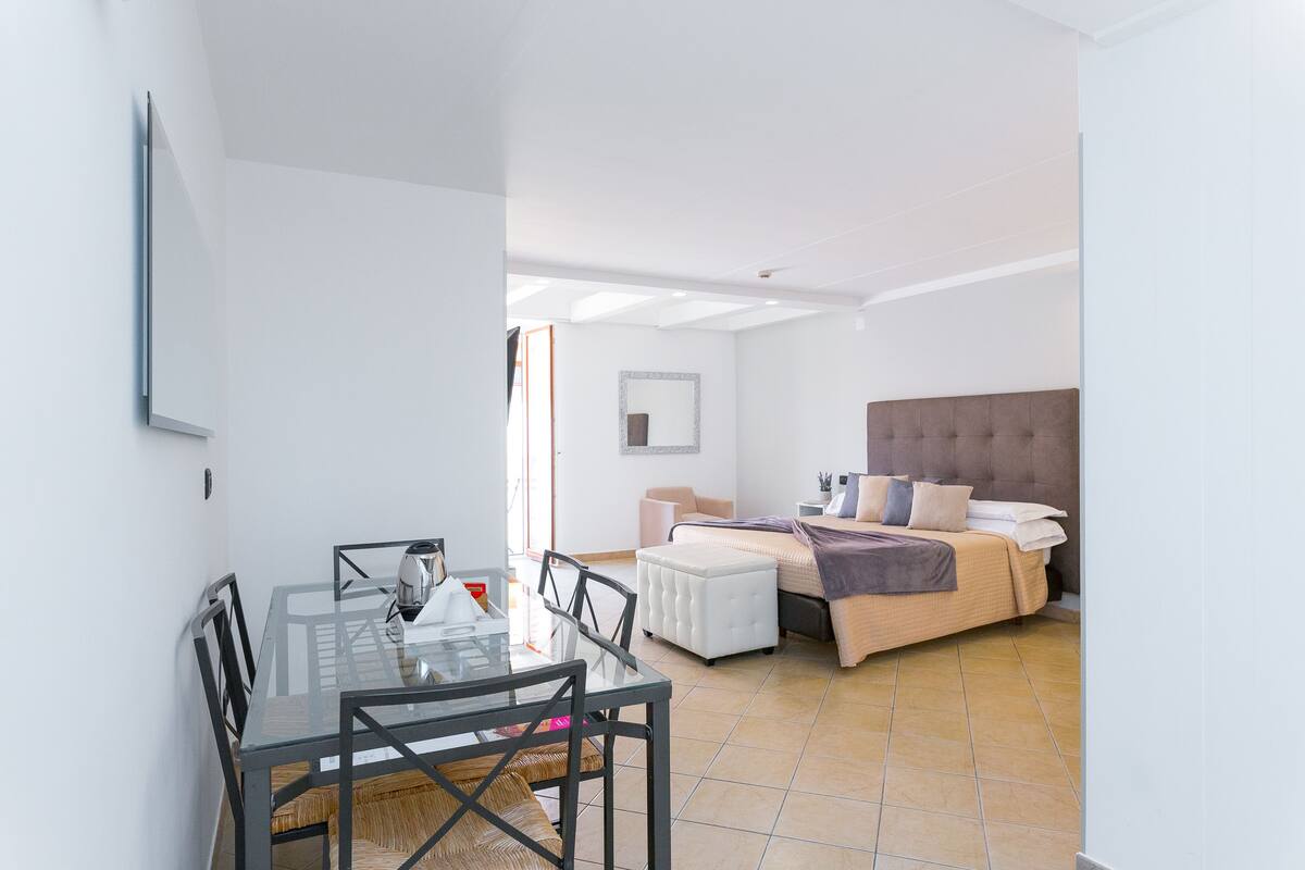 A modern room with a glass dining table, white walls, a bed with a headboard, and a small window.