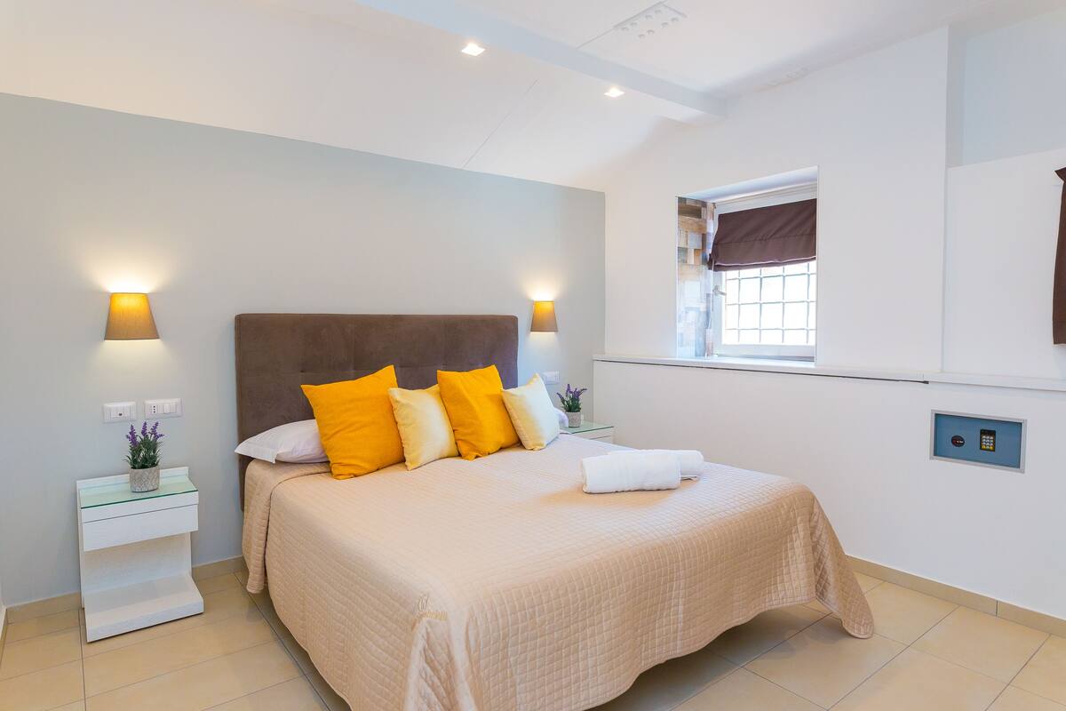 A neatly arranged bedroom with a bed, bedside lamps, and a window with curtains.