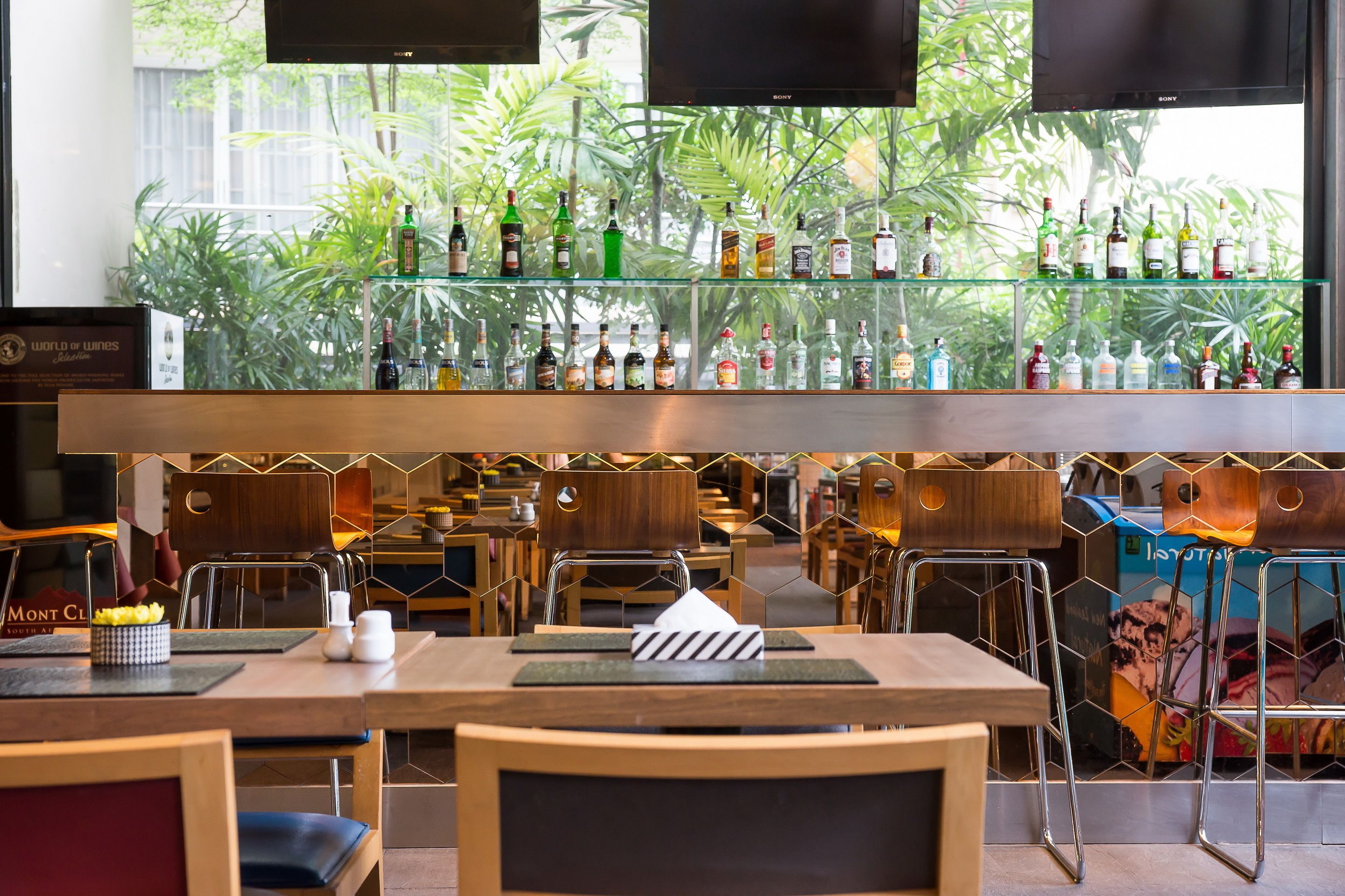 Een bar met een glazen plank waarop diverse flessen staan, houten stoelen en tafels met onderzetters en servetten.