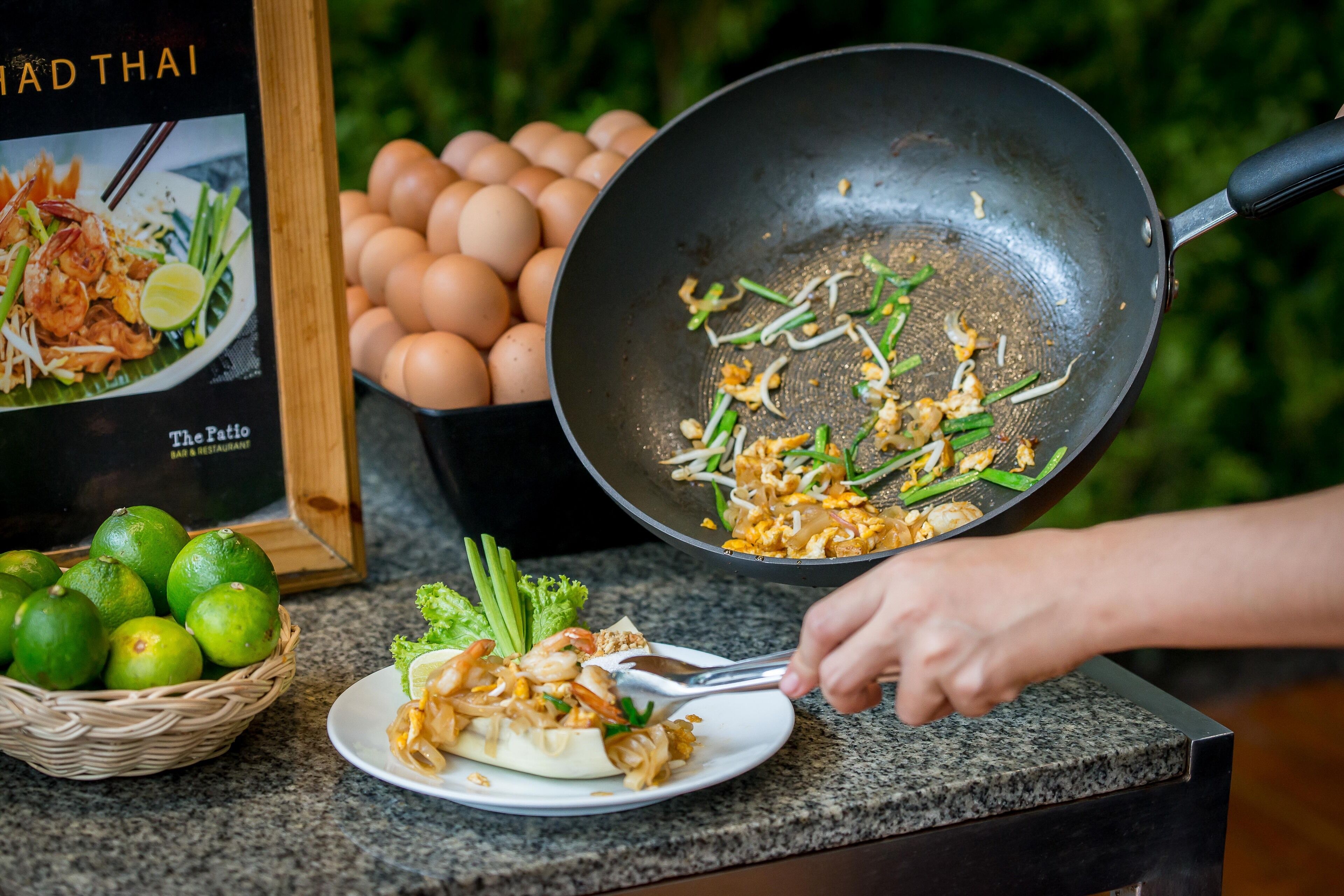 Eine Person kocht Pad Thai in einem Wok, dazu Eier und Limetten.