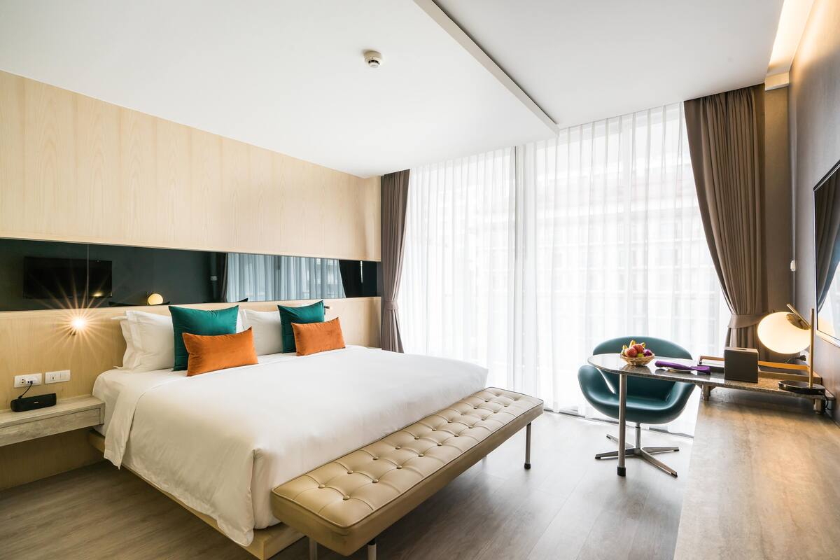 Ein modernes Hotelzimmer mit einem großen Bett, einer Bank, einem Schreibtisch mit einem Laptop und einem Sessel.