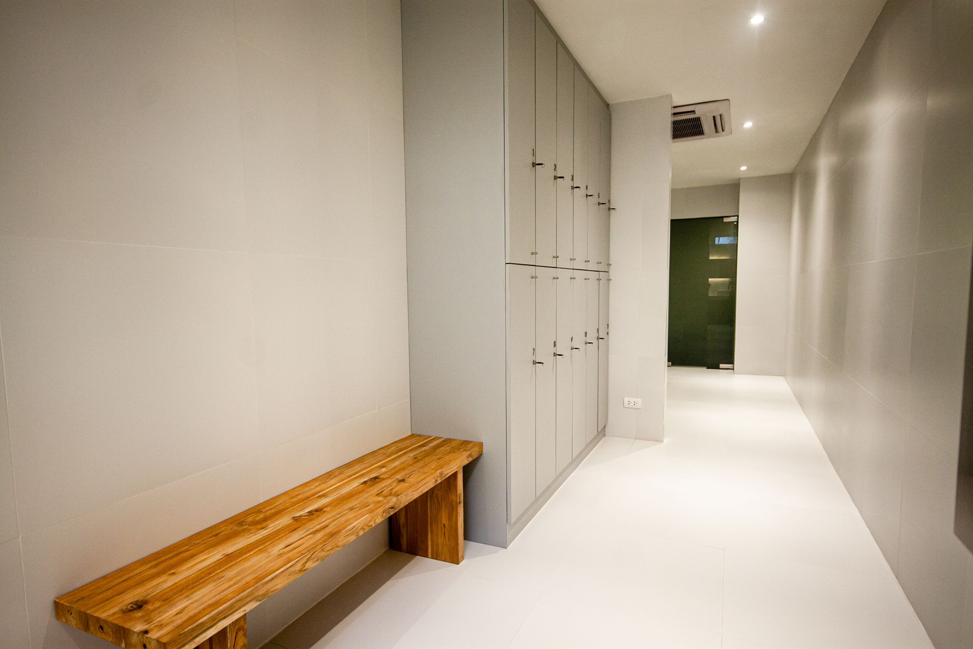 Un couloir avec un banc en bois, des casiers gris et un sol carrelé de carreaux blancs.