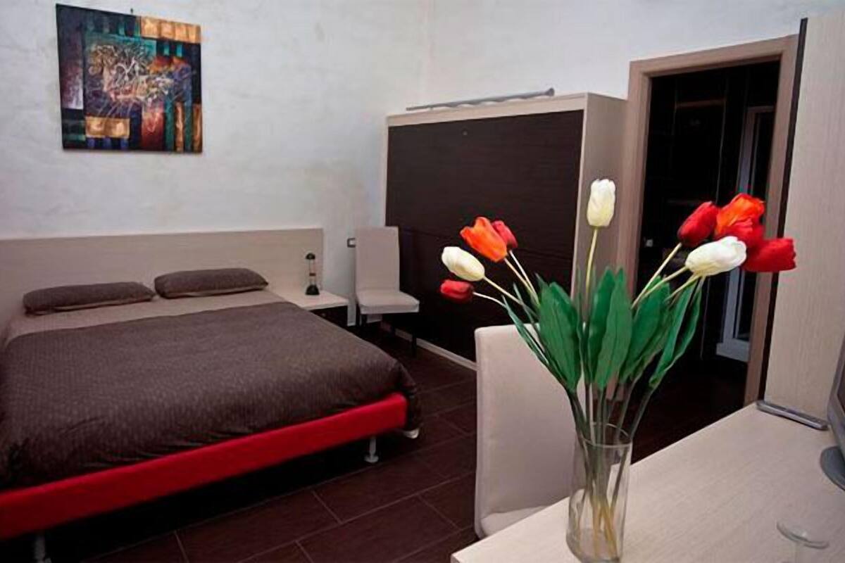 Ein Schlafzimmer mit Bett, Schreibtisch, Stuhl, Blumen in einer Vase und einem Bild an der Wand.