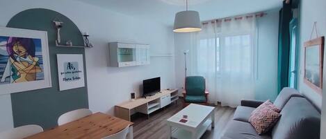 A modern living room with a wooden dining table, white chairs, a grey sofa, and a TV.