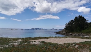 A coastal landscape with a sandy beach, a rocky shoreline, and a clear blue sky with scattered clouds.